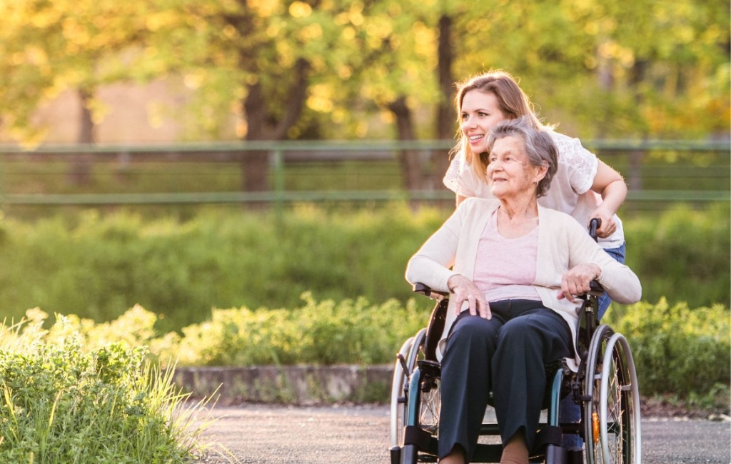 A smiling caregiver provides home care in Hallandale Beach, joyfully pushing an elderly woman in a wheelchair through a sunlit park surrounded by greenery.