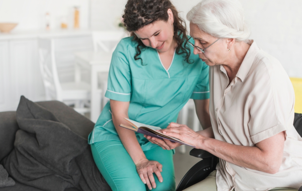 Miramar A caregiver in scrubs sitting beside a senior woman, both smiling and reading a book together in a cozy living space, representing compassionate home care in Miramar.